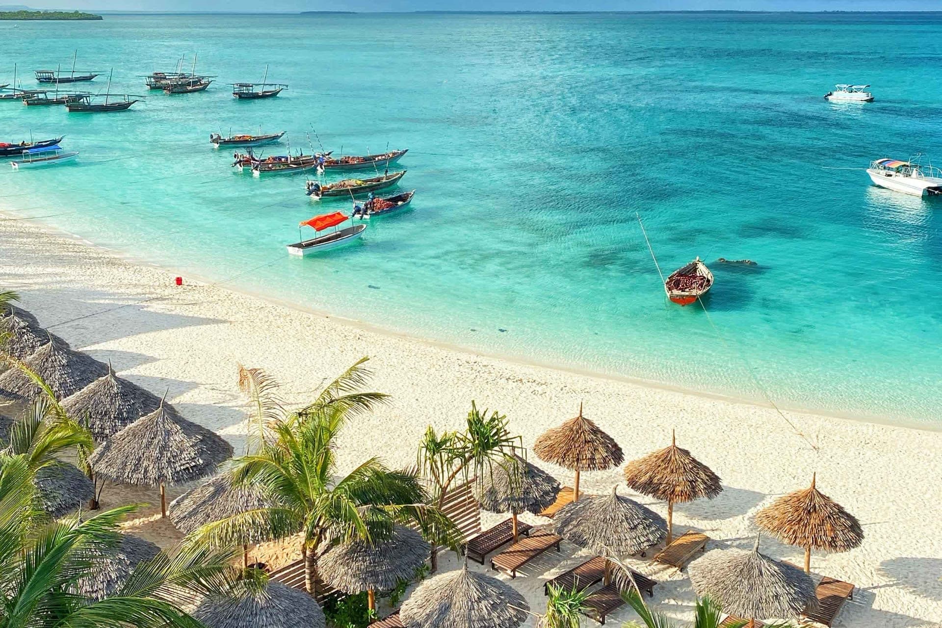 Thatched beach loungers and dhow boats anchored in turquoise water off Zanzibar